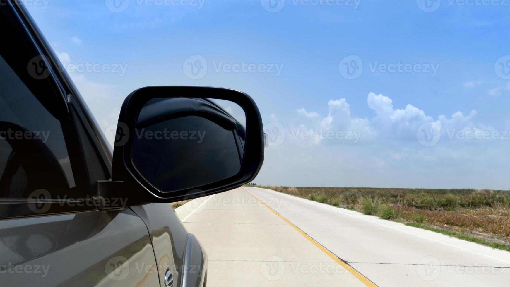 Beside of gray car driving on concrete road. Can view mirror wing of car. Beside with slope of concrete road and blurred of open space under blue sky. photo