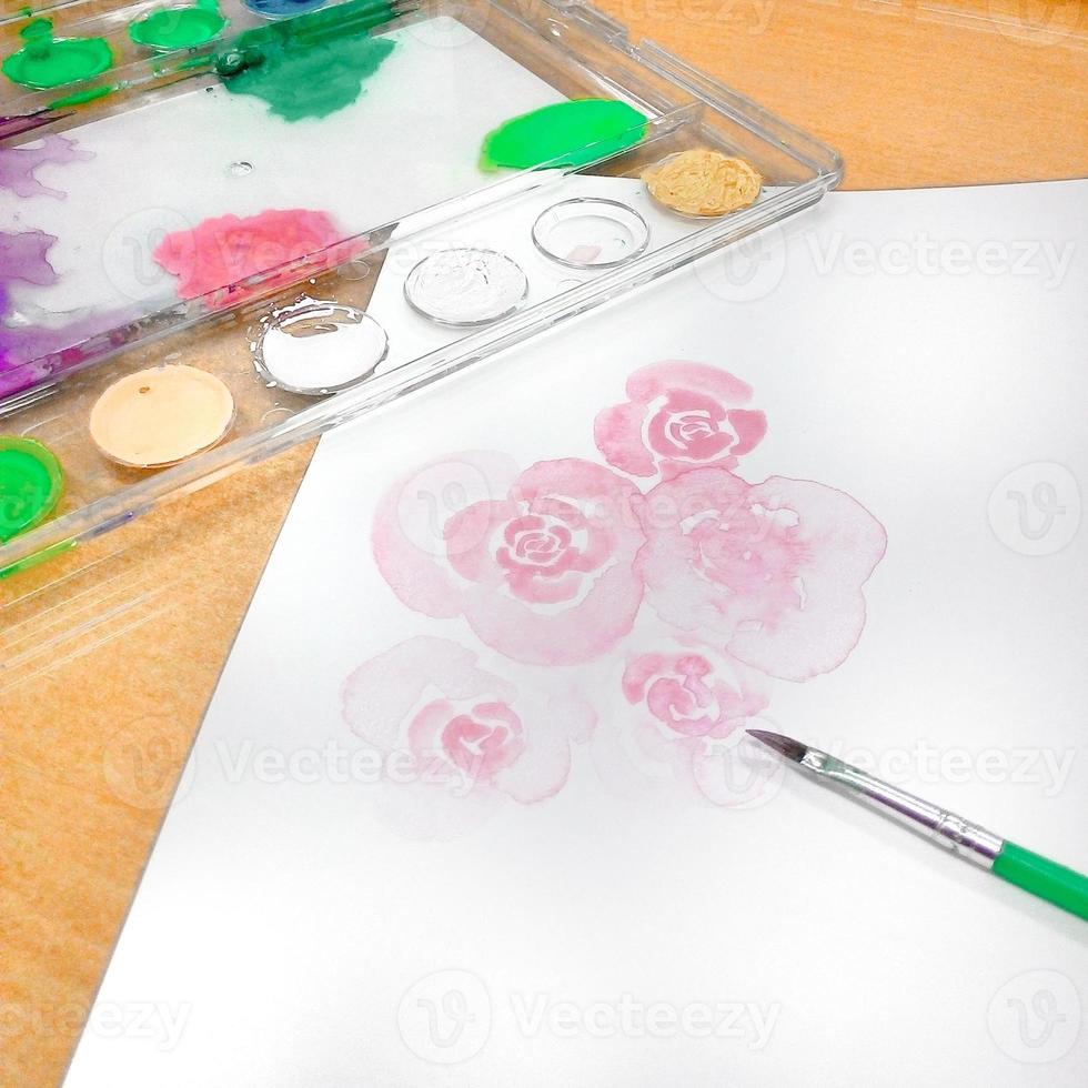 Watercolor paints and brushes on a white sheet of paper with a rose drawing. photo