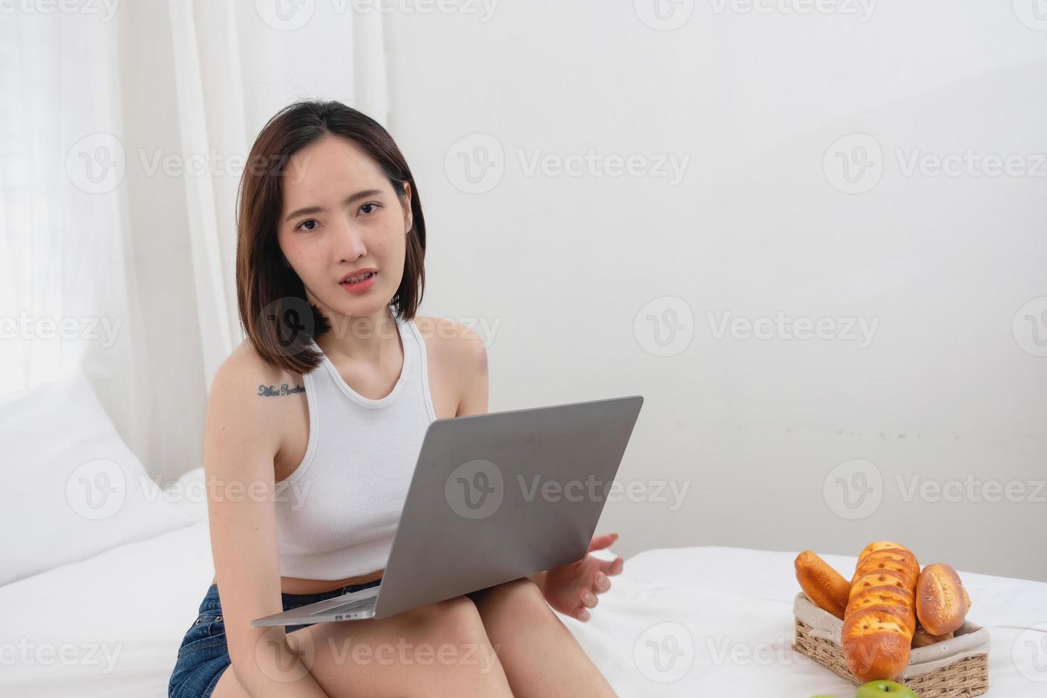 Portrait of white Asian woman with a tattoo sitting on the bed relaxing and playing laptop on weekend There was bread and fruit and red apples on a white bed to eat. holiday concept photo