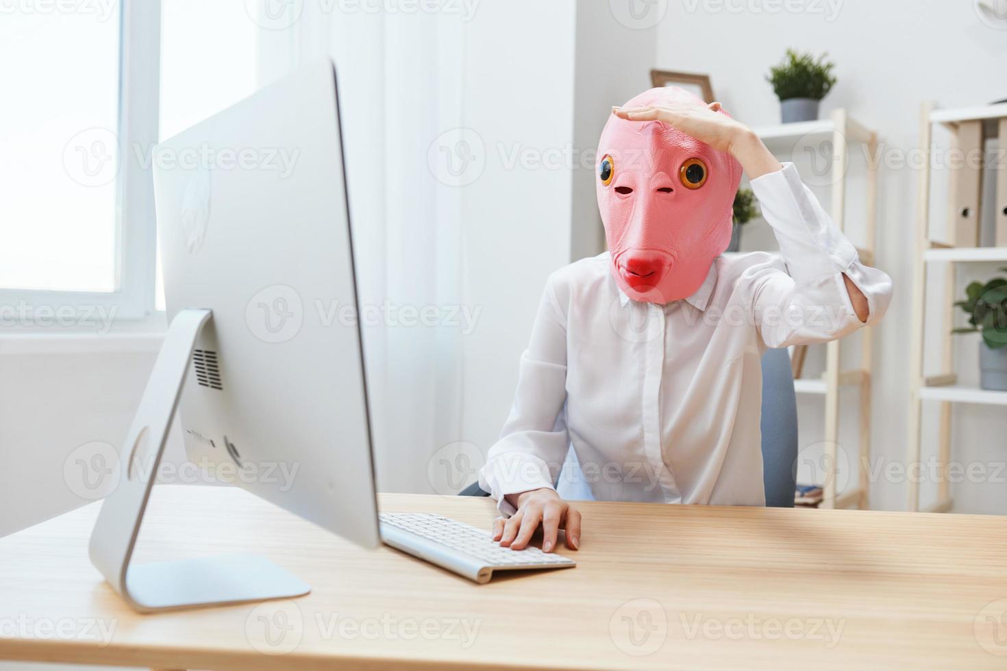 Funny crazy businesswoman worker freelancer in pink fish mask work on computer online pondering or making decision holds hand over eyes and looks into distance in light modern office. Copy space photo