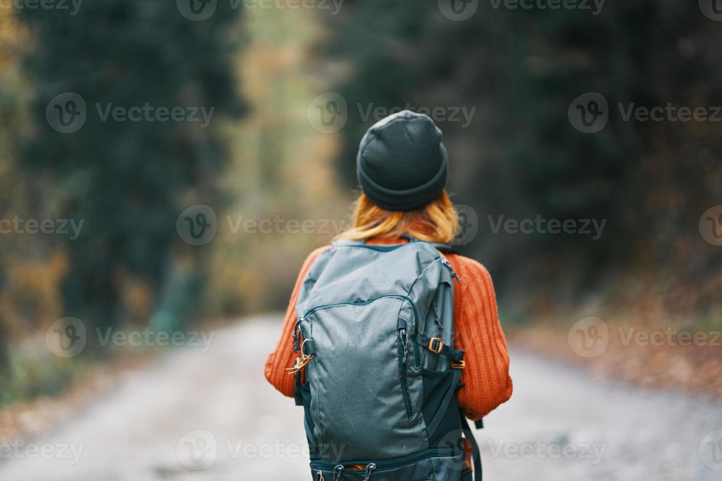 woman with a backpack in a hat and an orange sweater on the road in the autumn forest back view photo