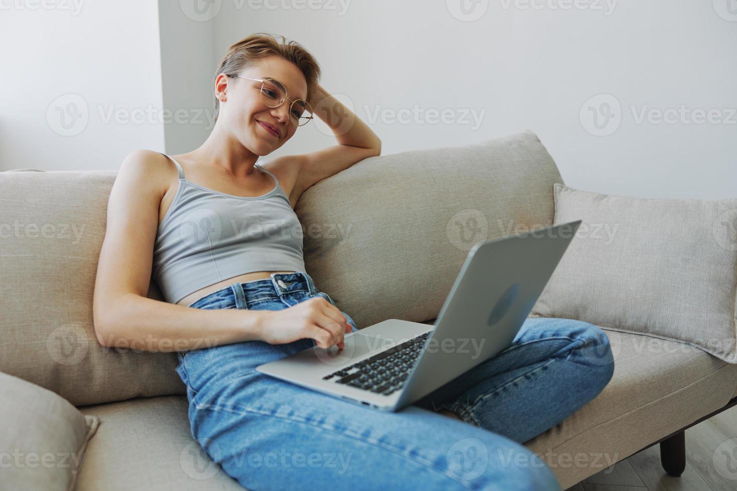 Teenage girl freelancer with laptop sitting on couch at home smiling in home clothes and glasses with short haircut, lifestyle with no filters, free copy space photo
