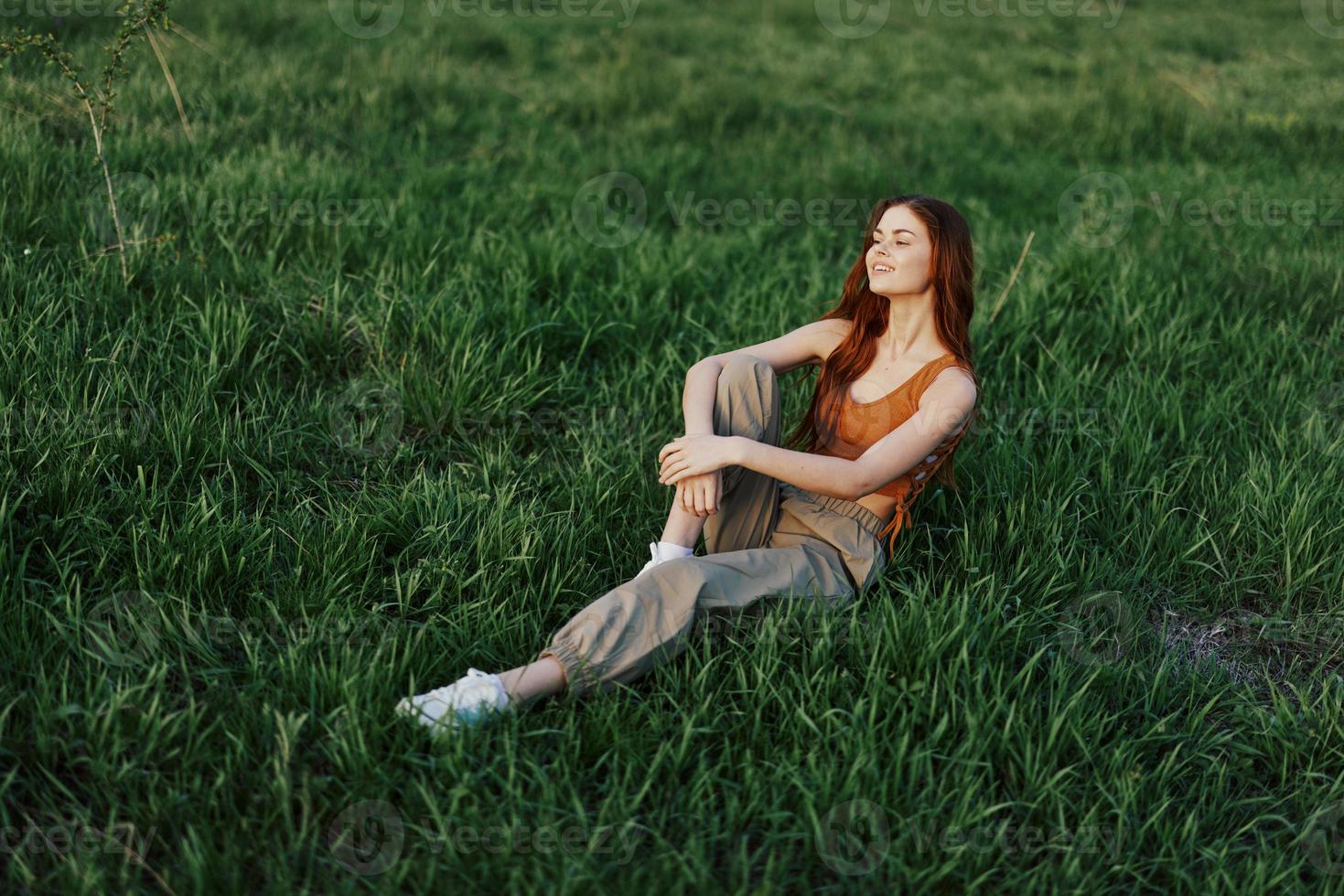 The redheaded woman sits in the park on the green grass wearing an orange top, green pants, and sneakers and looks out at the setting summer sun photo