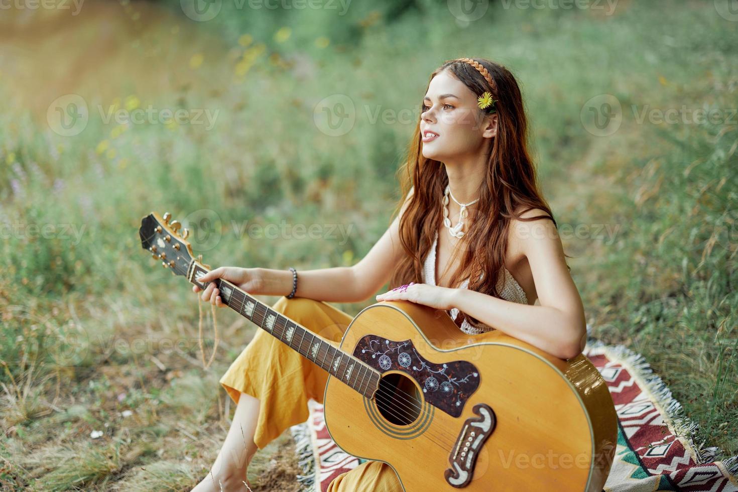 hippie mujer eco simpático jugando guitarra sonrisas y canto canciones en naturaleza sentado en un tartán por el lago en el noche en el puesta de sol luz de sol. un estilo de vida en armonía con el cuerpo y naturaleza foto