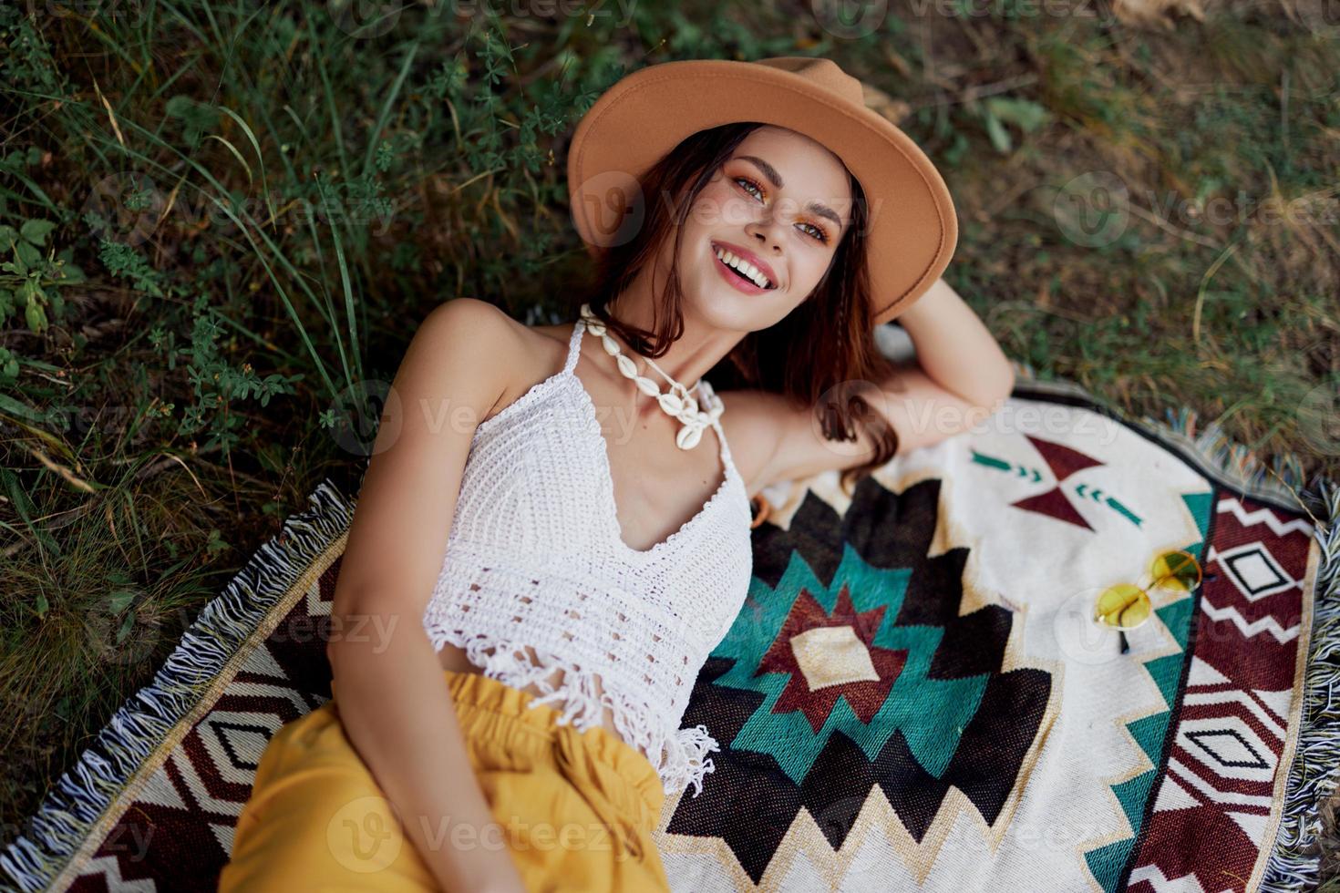 A woman in eco-friendly clothing in a hippie look is lying on a colored plaid smiling and looking at an autumn sunset in nature in the park. Lifestyle on a journey of harmony with the world photo