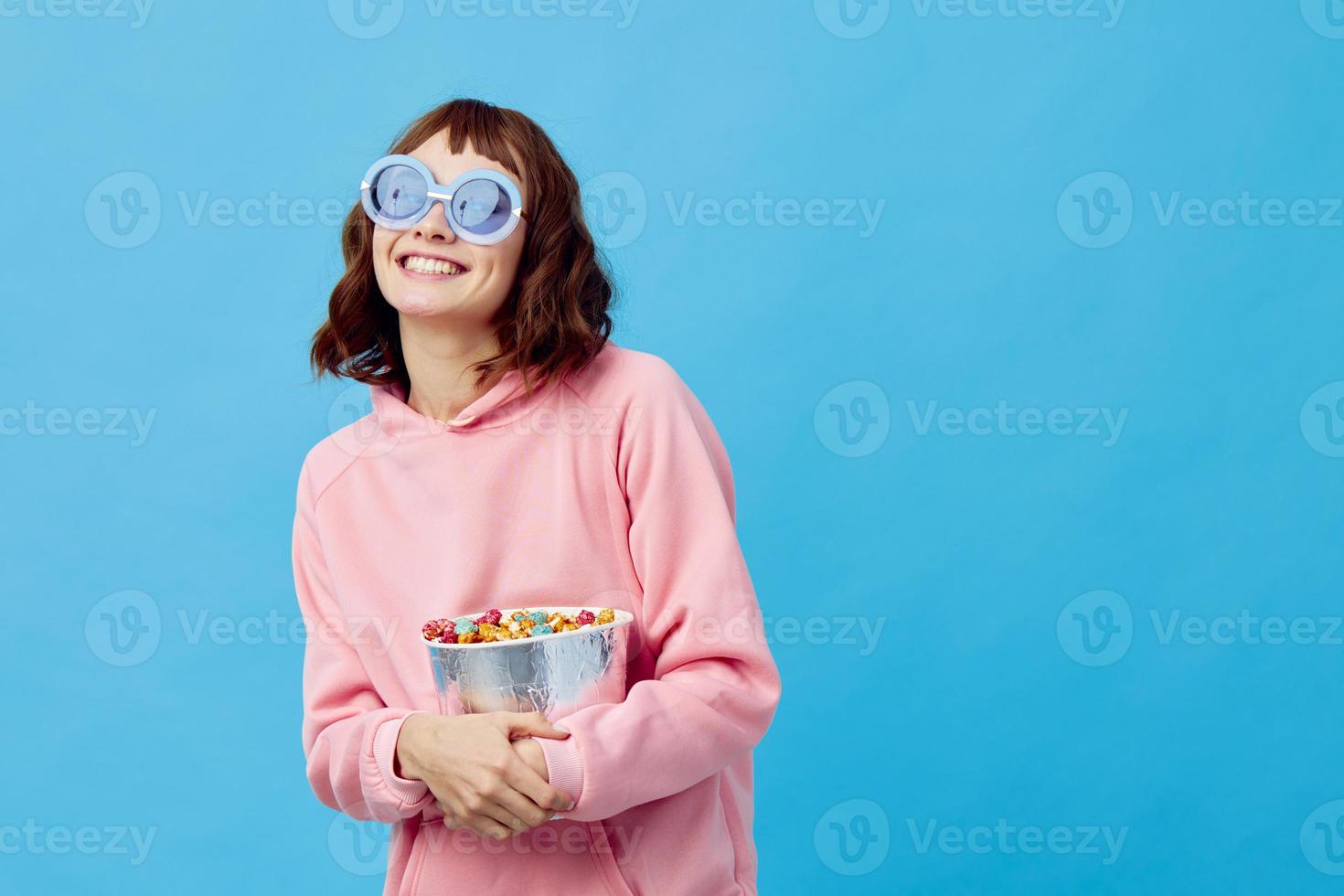 noche película concepto. encantador sonriente linda pelirrojo dama en rosado capucha Gafas de sol con palomitas de maiz posando aislado en azul estudio antecedentes. Copiar espacio bandera. Moda cine. entretenimiento oferta foto