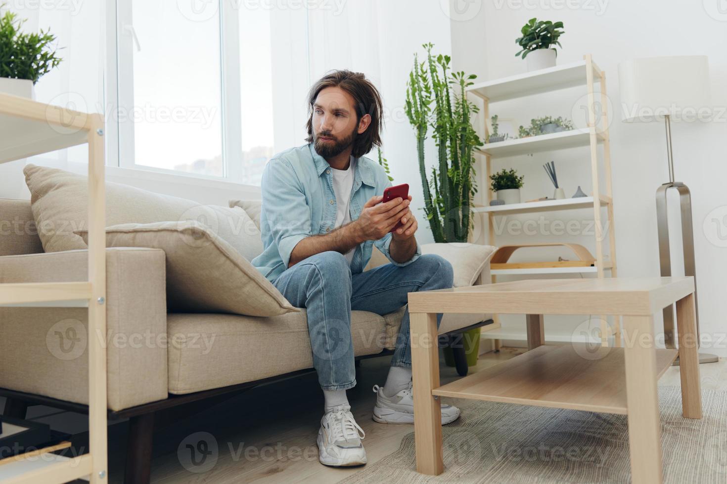 A man with a beard sits on the couch during the day and smiles at home and looks at his phone relaxing on his day off, life online on his phone texting with friends photo