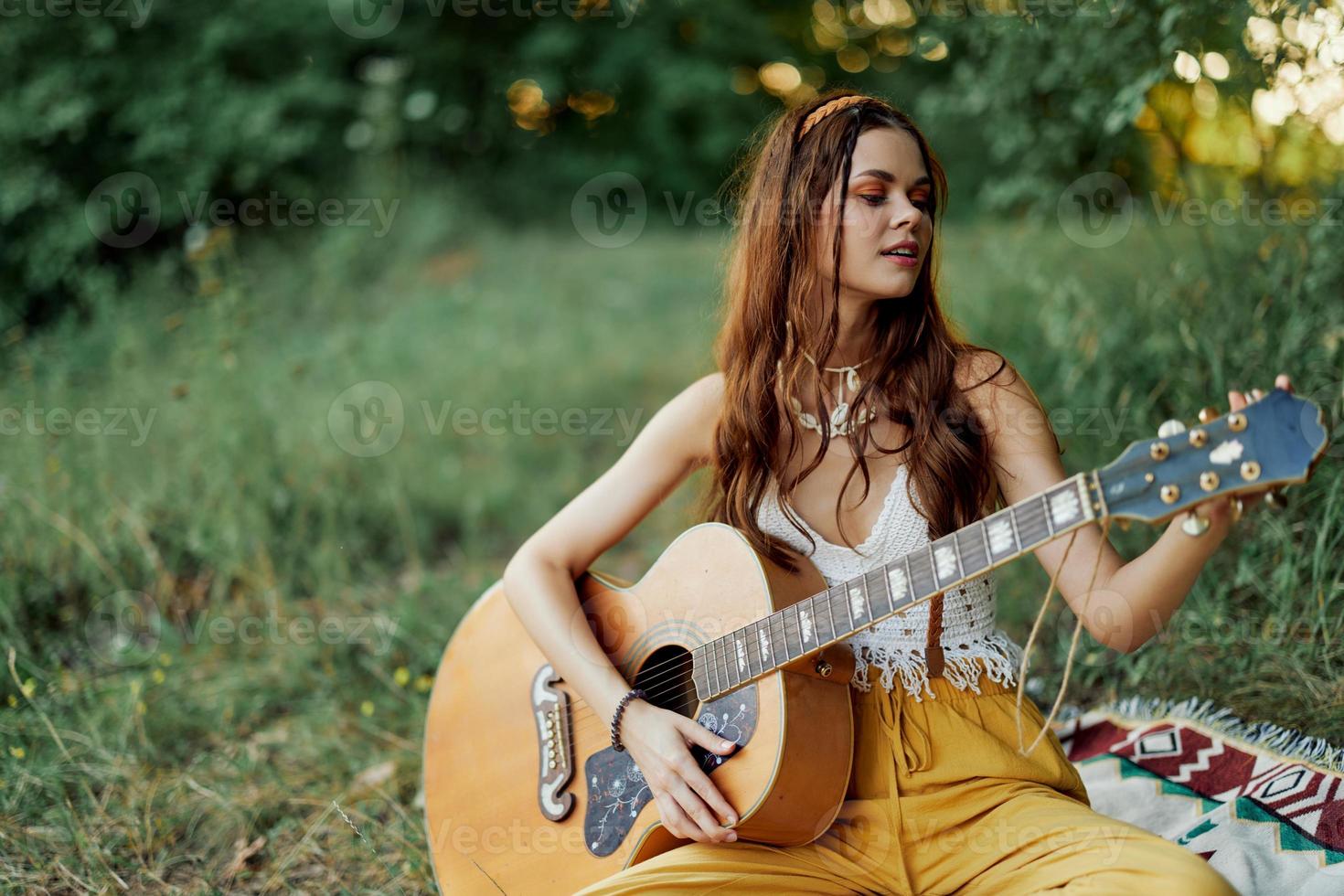 niña hippie mujer jugando guitarra en Respetuoso del medio ambiente ropa sentado en el suelo fuera de en naturaleza en el otoño acecho el puesta de sol foto
