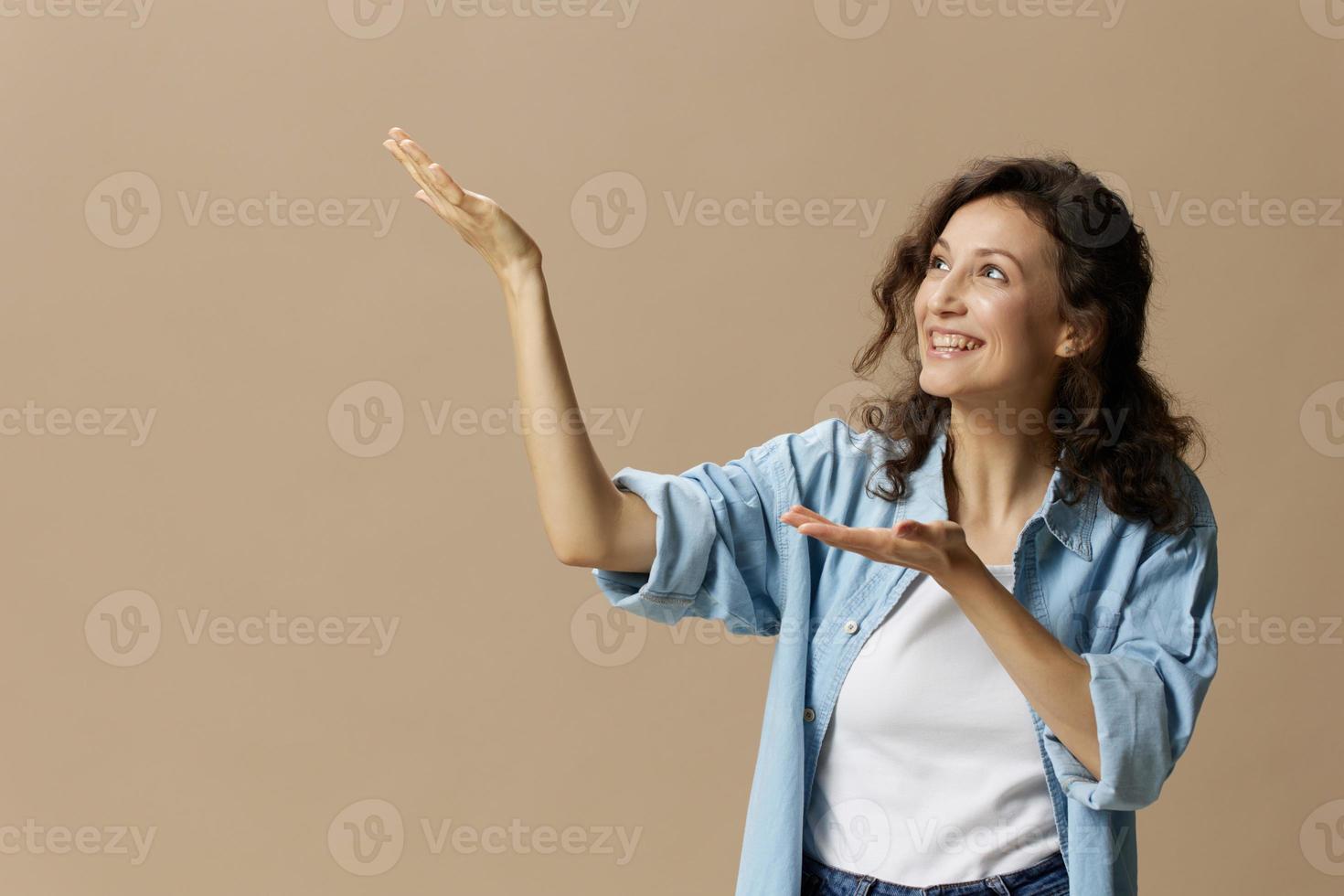 Cheerful happy curly beautiful lady in jeans casual shirt point both hands looks up at free place for ad posing isolated on over beige pastel background. People Lifestyle emotions concept. Copy space photo