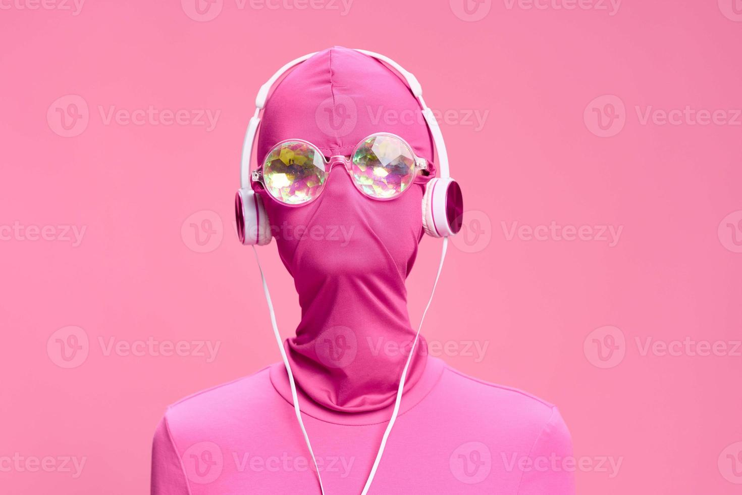 Art portrait of a woman wearing a pink full-face burglar mask with glowing round glasses wearing pink clothes with pink headphones on a pink background looking into the camera photo