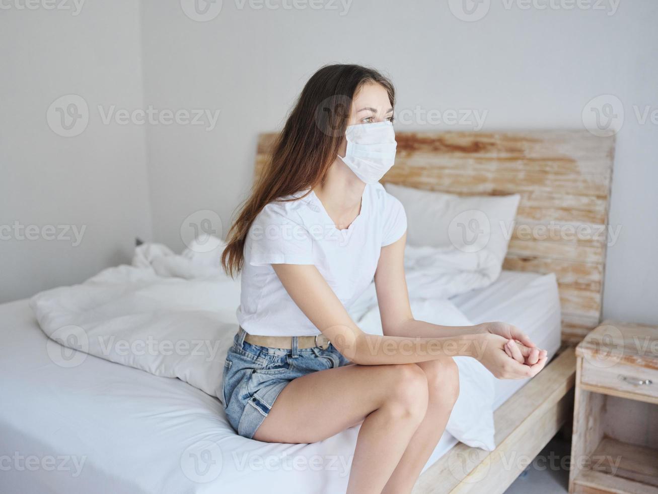side view of pretty woman in medical mask sitting on bed in isolated room photo