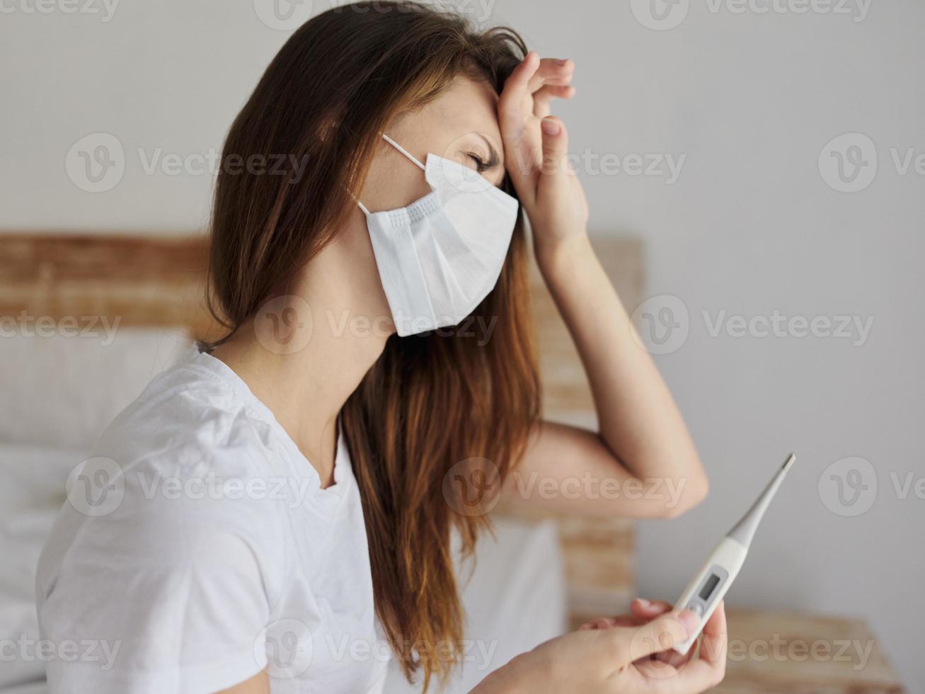 woman holding hand on forehead thermometer in hands medical mask checking temperature photo