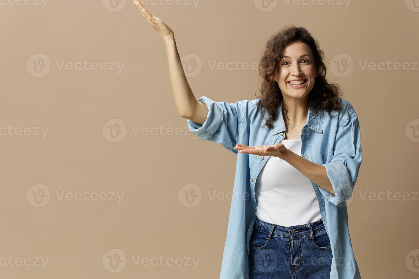 Cheerful happy curly beautiful female in jeans casual shirt point both hands up at free place for ad posing isolated on over beige pastel background. People Lifestyle emotions concept. Copy space photo