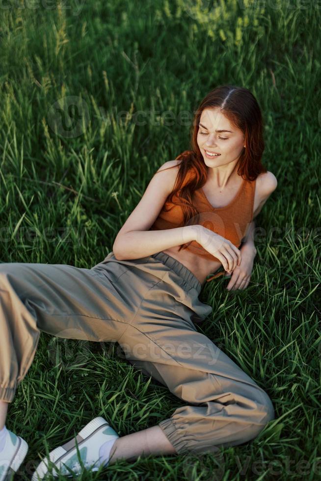 un mujer acostado en el verde césped en verde pantalones y un naranja arriba, sonriente en el puesta de sol verano Dom en naturaleza foto