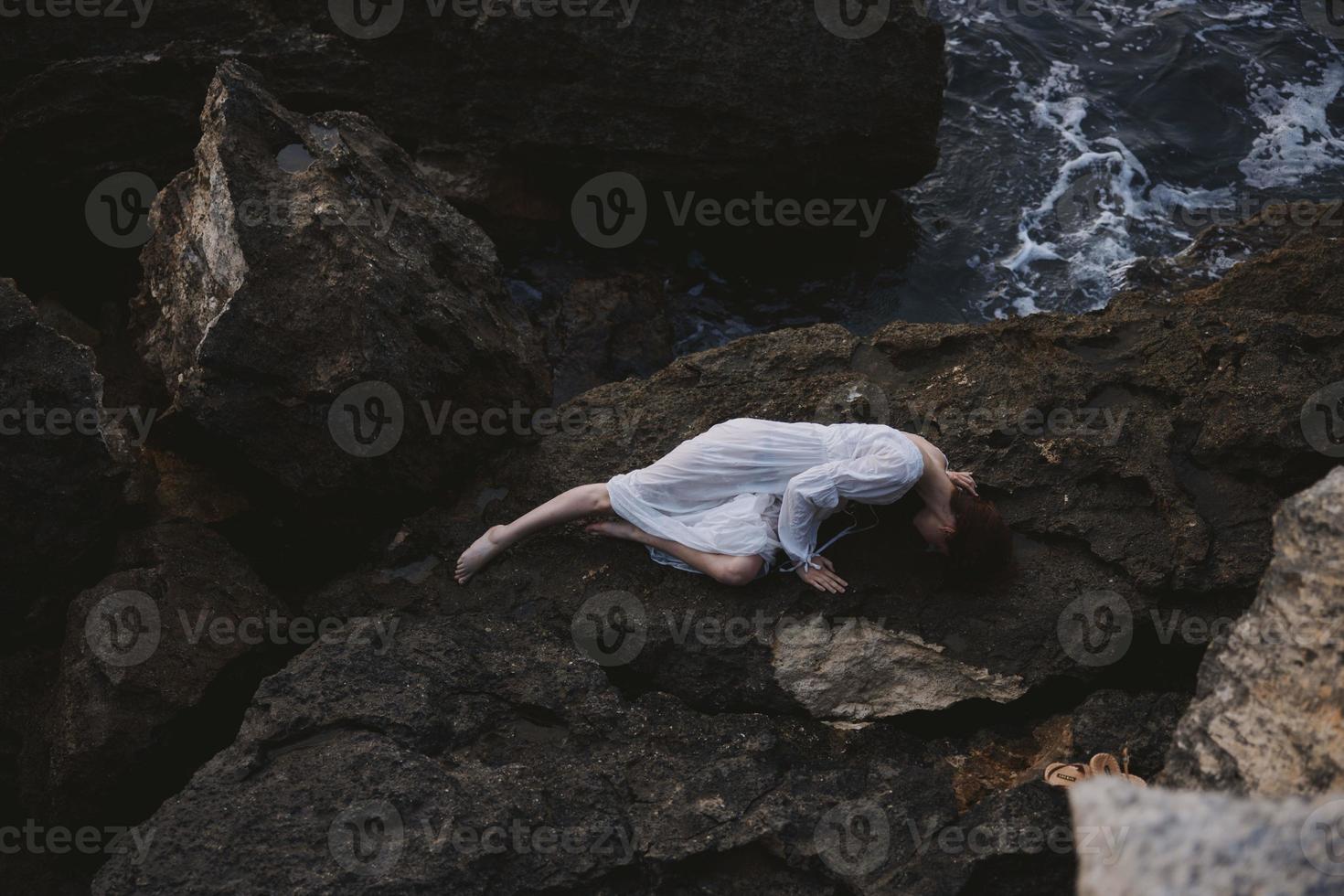 descalzo mujer en un aislado Mancha en un salvaje rocoso costa en un blanco vestir ver desde encima foto