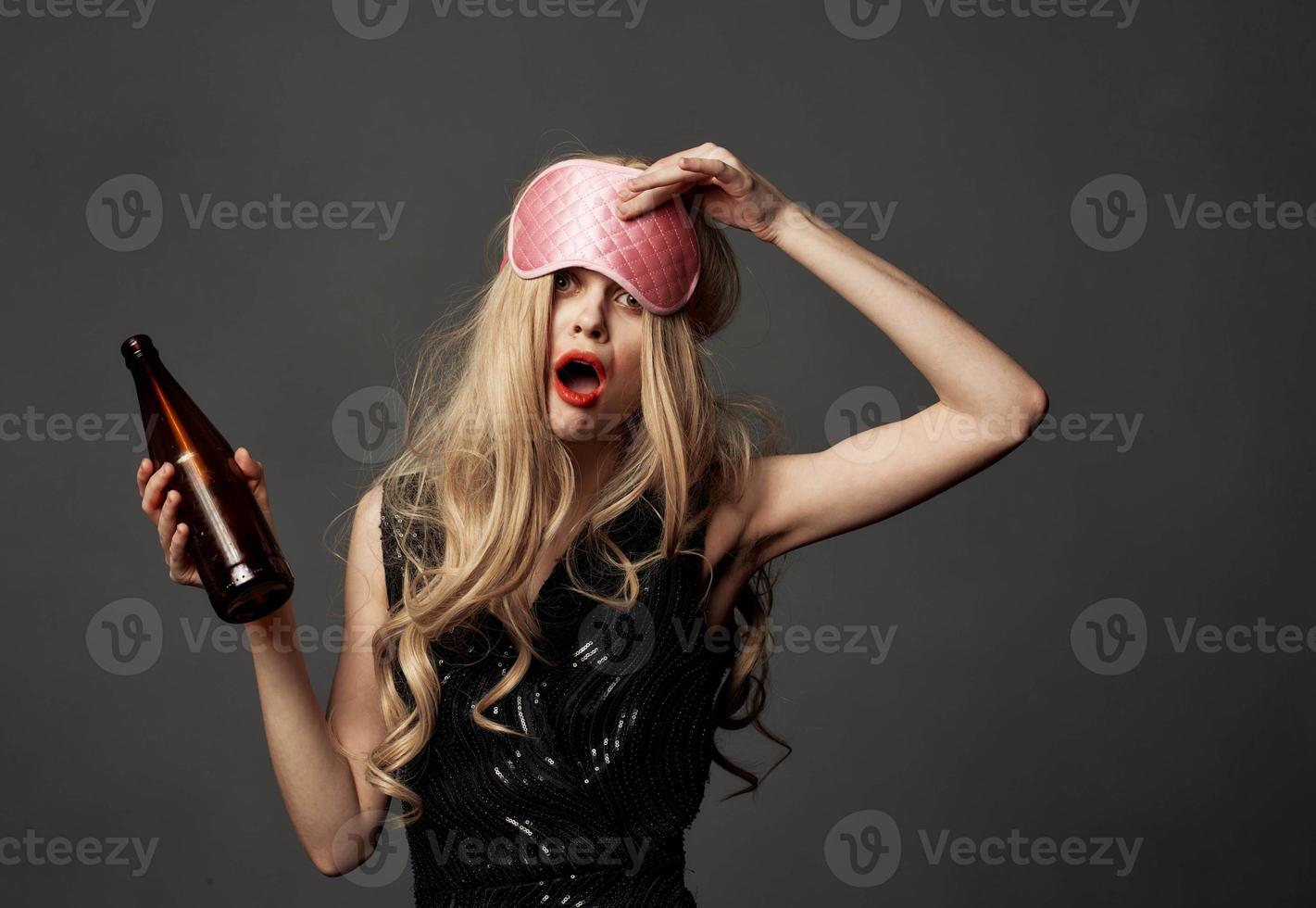 Drunk woman with a bottle of alcohol in her hand on a gray background and a mask for sleeping photo