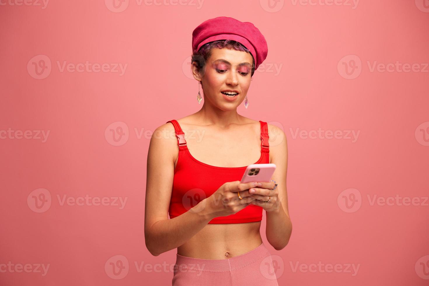 Young woman blogger with colored pink hair and short haircut flipping through the phone screen and typing a message with a smile and surprise in stylish clothes on a pink background photo