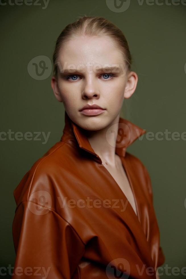 Stylish serious young blonde girl with blue eyes with evening makeup in a leather dress looks at the camera posing isolated. Studio portrait on a green background. Fashion offer photo