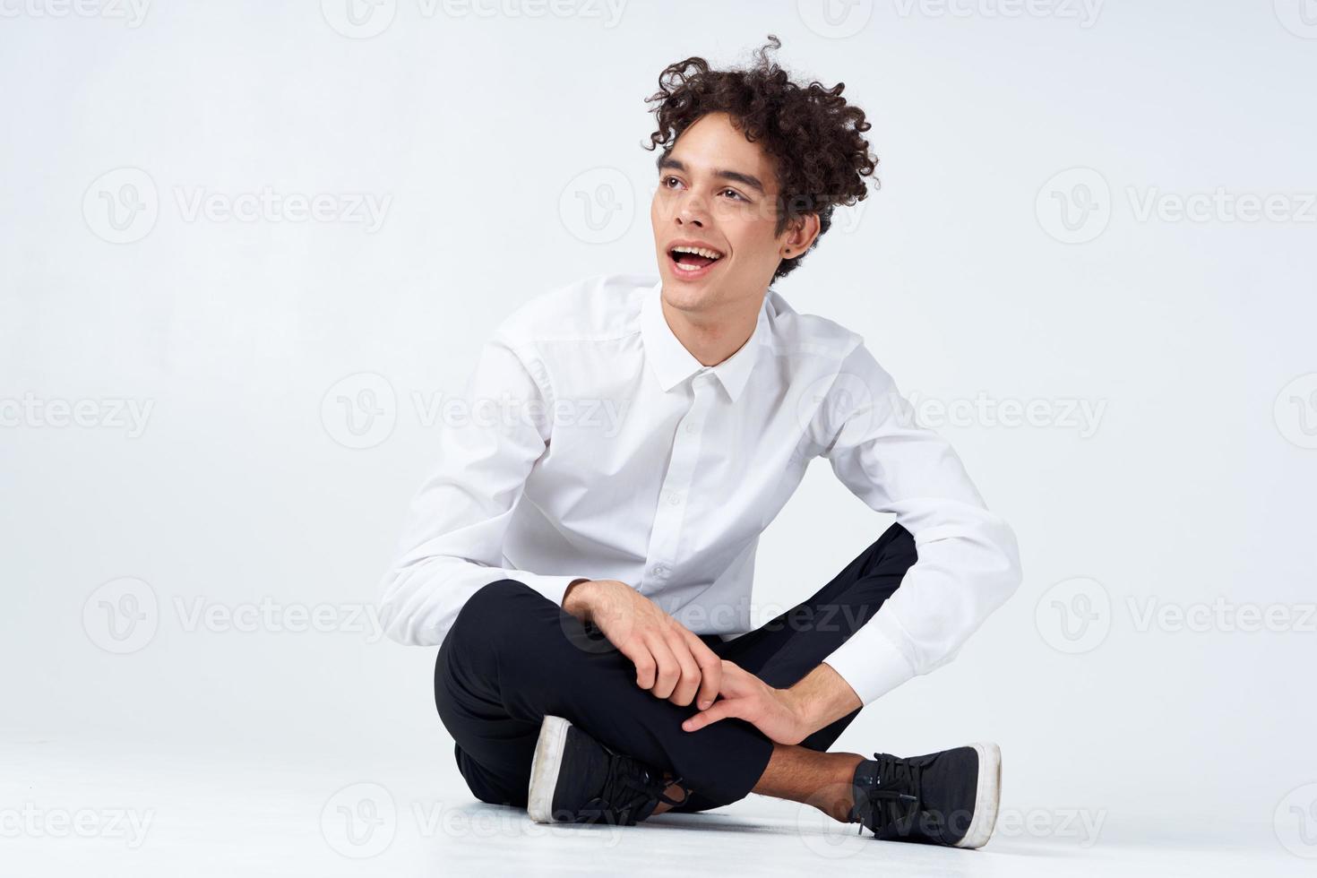 a guy in trousers in sneakers and a shirt sits on the floor and gestures with his hands photo