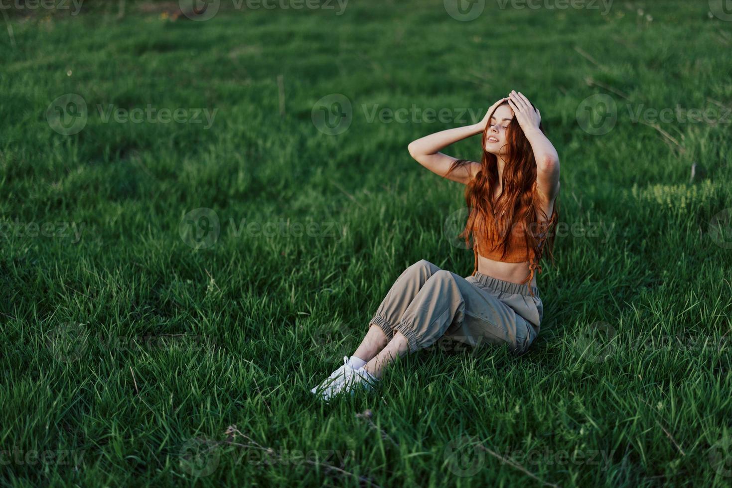 A red-haired woman with long, windswept hair sits outdoors on the grass in the park and smiles, the sunset light illuminating her face. The concept of harmony with nature outdoors photo