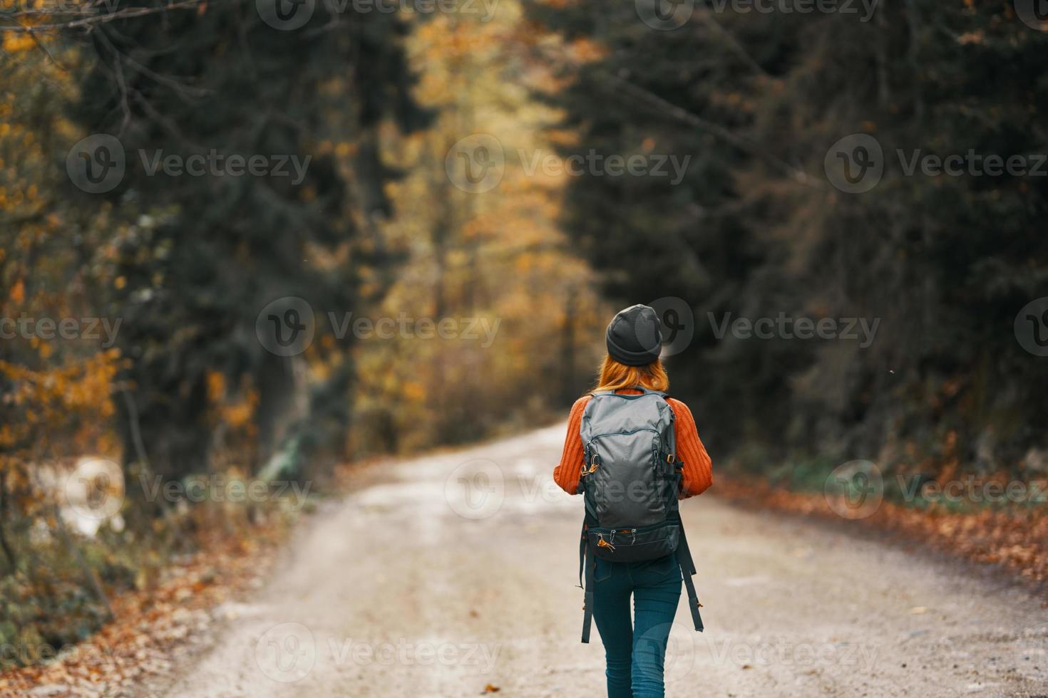 woman with a backpack in a hat and an orange sweater on the road in the autumn forest photo