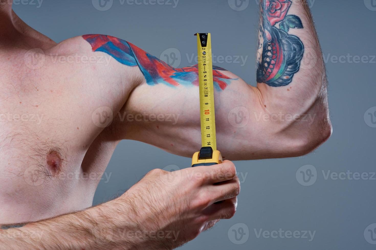 guy measures arm muscles with centimeters on a gray background and a multicolored tattoo bodybuilder fitness photo