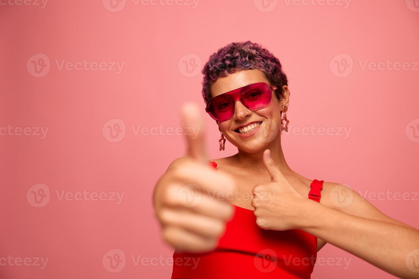 Fashion portrait of a woman with a short haircut in colored sunglasses with unusual accessories with earrings smiling on a pink bright background showing thumbs up at the camera photo