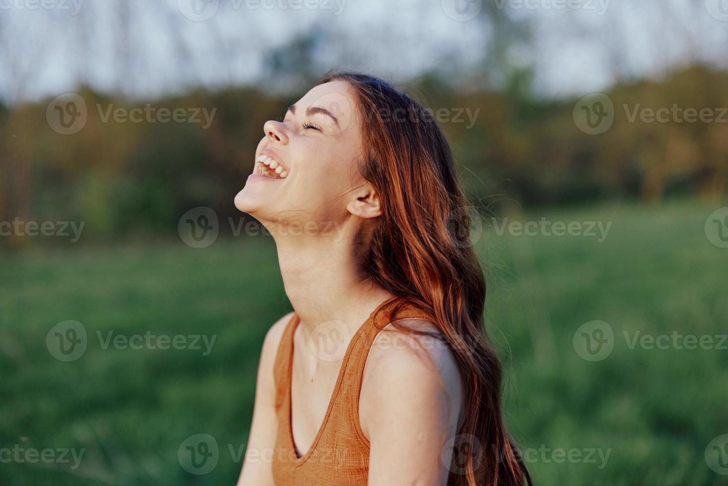 un joven mujer riendo y sonriente alegremente en naturaleza en el parque con el puesta de sol Encendiendo esclarecedor su largo rojo pelo foto