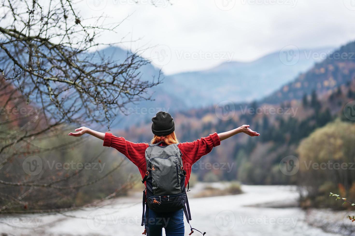 a traveler in a hat, sweater and trousers gestures with her hands on the river bank in the mountains photo