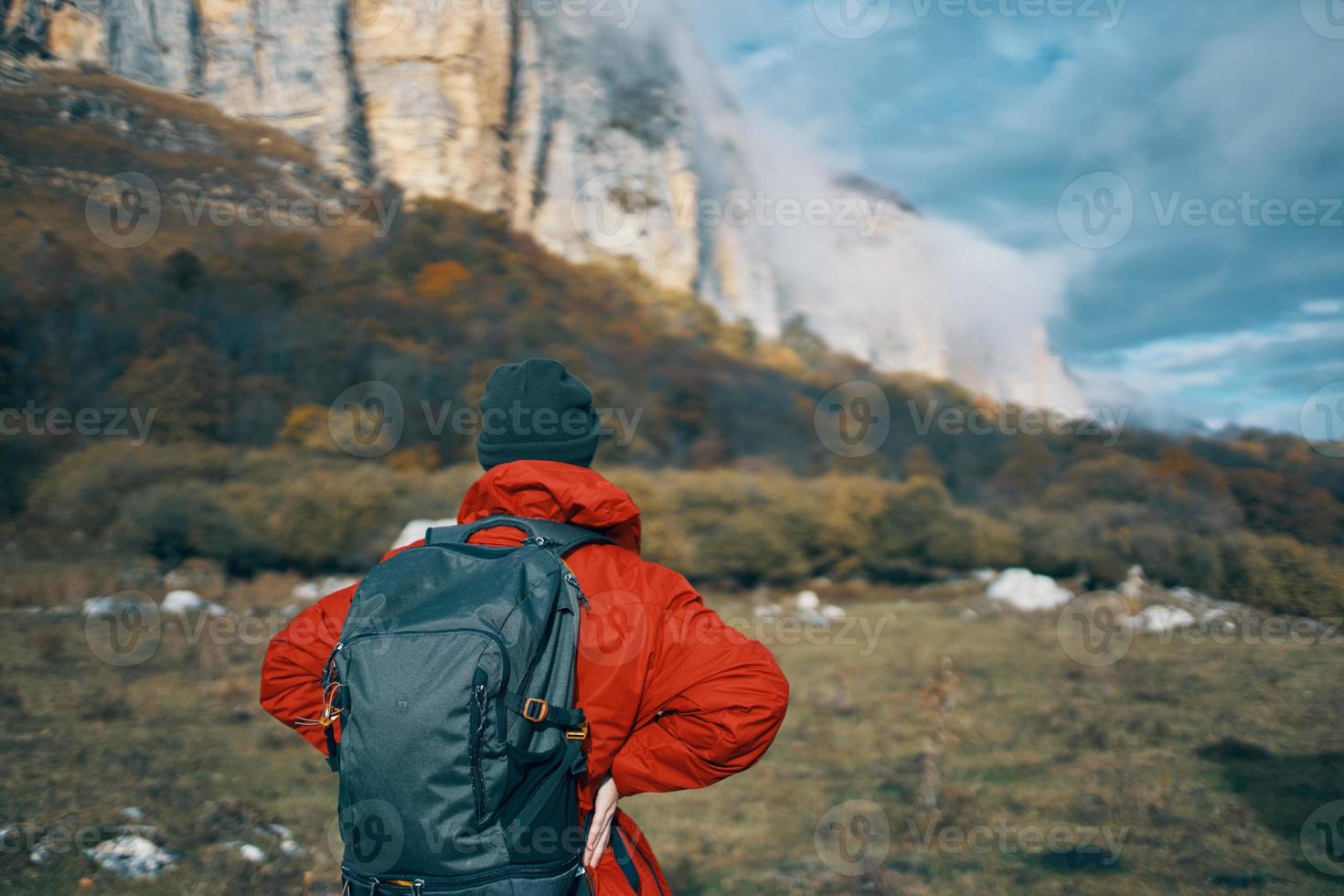 a woman in a red jacket with a backpack and in a warm hat walks on the grass in the fall in the mountains photo