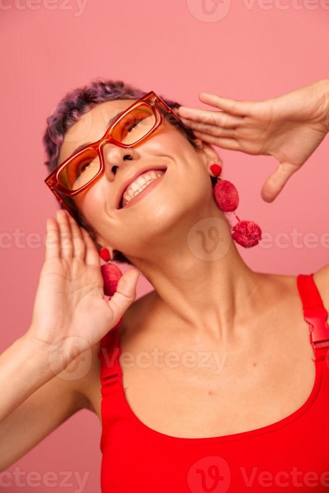 joven atlético mujer con un corto Corte de pelo y púrpura pelo en un rojo parte superior y rosado yoga polainas en Gafas de sol con un atlético figura sonrisas y bailes en un rosado antecedentes foto