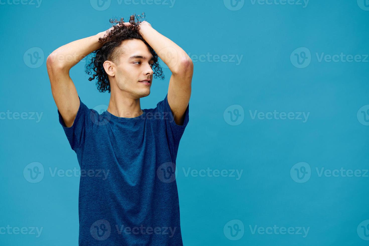 Positive joyful curly tanned Caucasian guy in basic t-shirt touches plays with hair curls posing isolated on over blue background. Lifestyle and Emotions concept. Good offer with copy space for ad photo