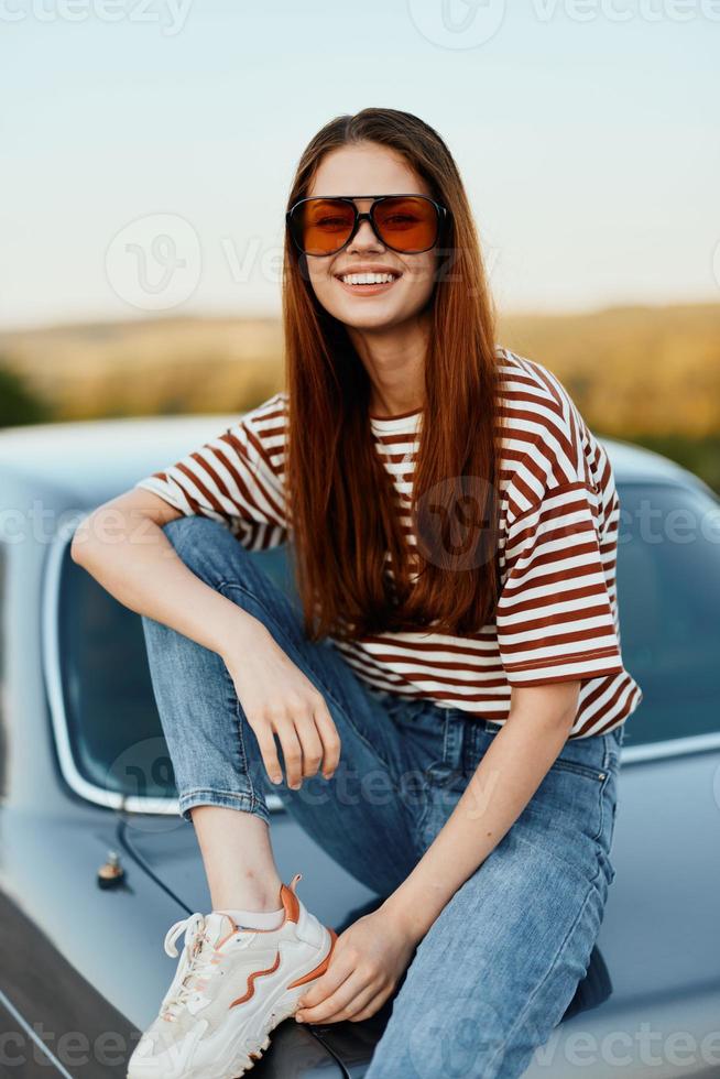 A fashionable woman smiles sweetly in stylish sunglasses, a striped t-shirt and jeans, sits on the trunk of a car and looks at the beautiful nature of autumn. Travel lifestyle photo