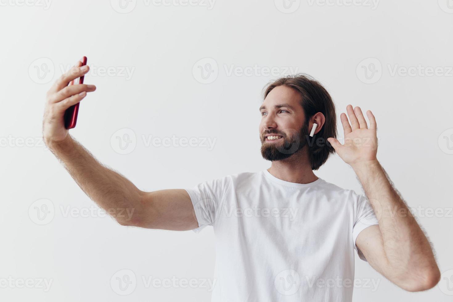 un masculino blogger grabando en video él mismo en su teléfono y chateando con personas en línea con un sonrisa en un blanco camiseta en contra un blanco pared foto