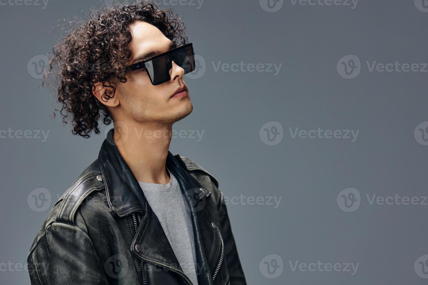 Stylish tanned curly man leather jacket mirror trendy eyewear looks aside posing isolated on gray studio background. Cool fashion offer. Huge Seasonal Sale New Collection concept. Copy space for ad photo