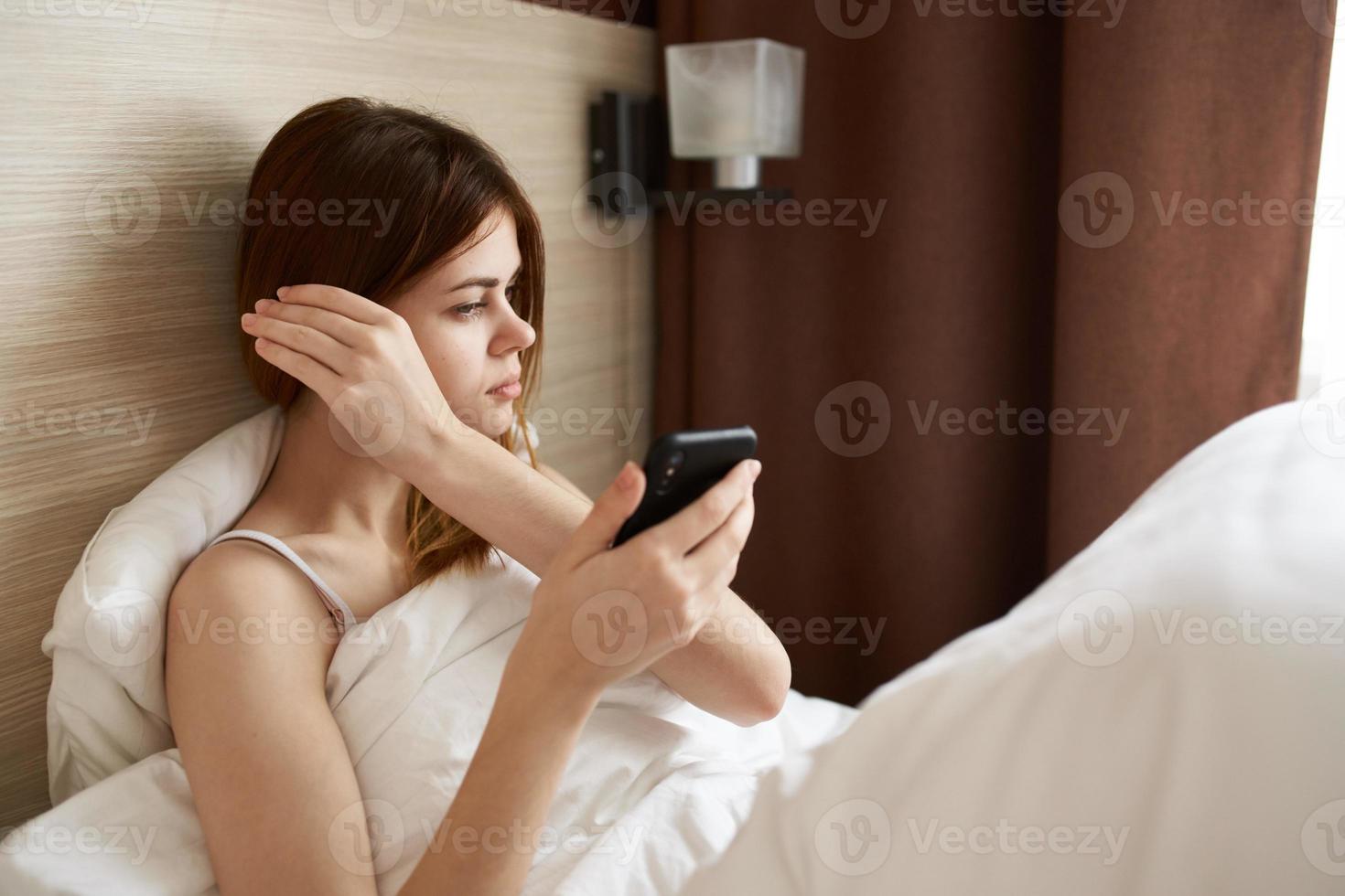 woman lies in bed under the covers with a mobile phone in her hand interior curtains design photo