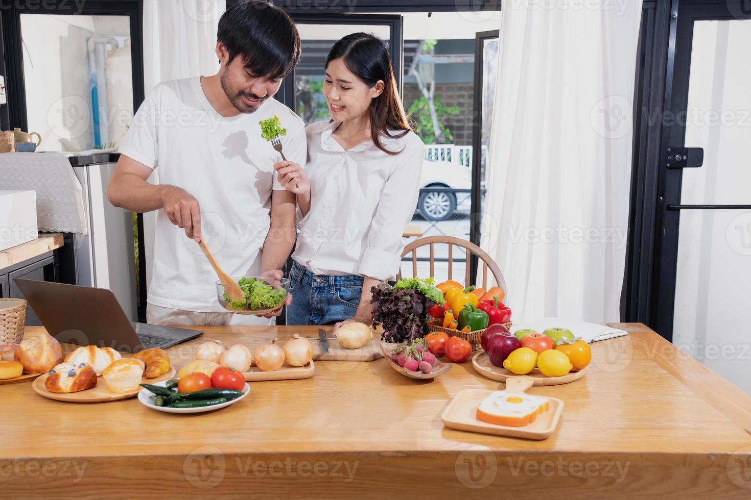 Young Asian couple cooking with fruits and vegetables and using laptop in the kitchen To cook food together within the family happily, family concept. photo