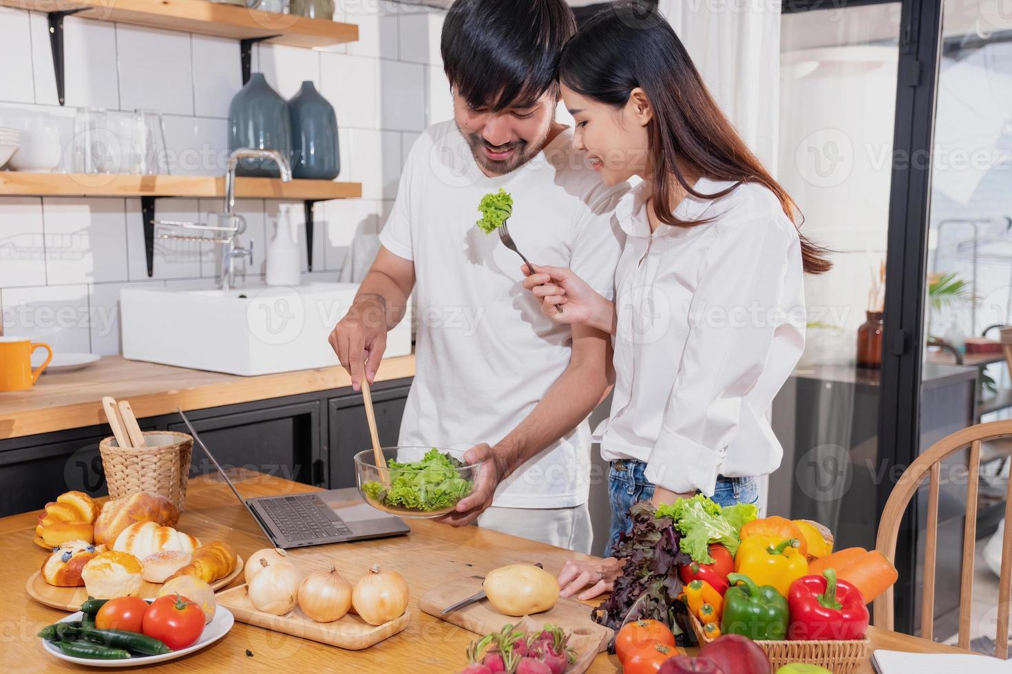 Young Asian couple cooking with fruits and vegetables and using laptop in the kitchen To cook food together within the family happily, family concept. photo