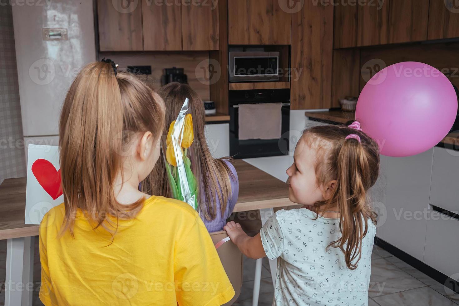 hijas felicitar su mamá en de la madre día, un tarjeta con un corazón, flores y un globo a hogar en el cocina. niños sorpresa su madre para el día festivo. foto