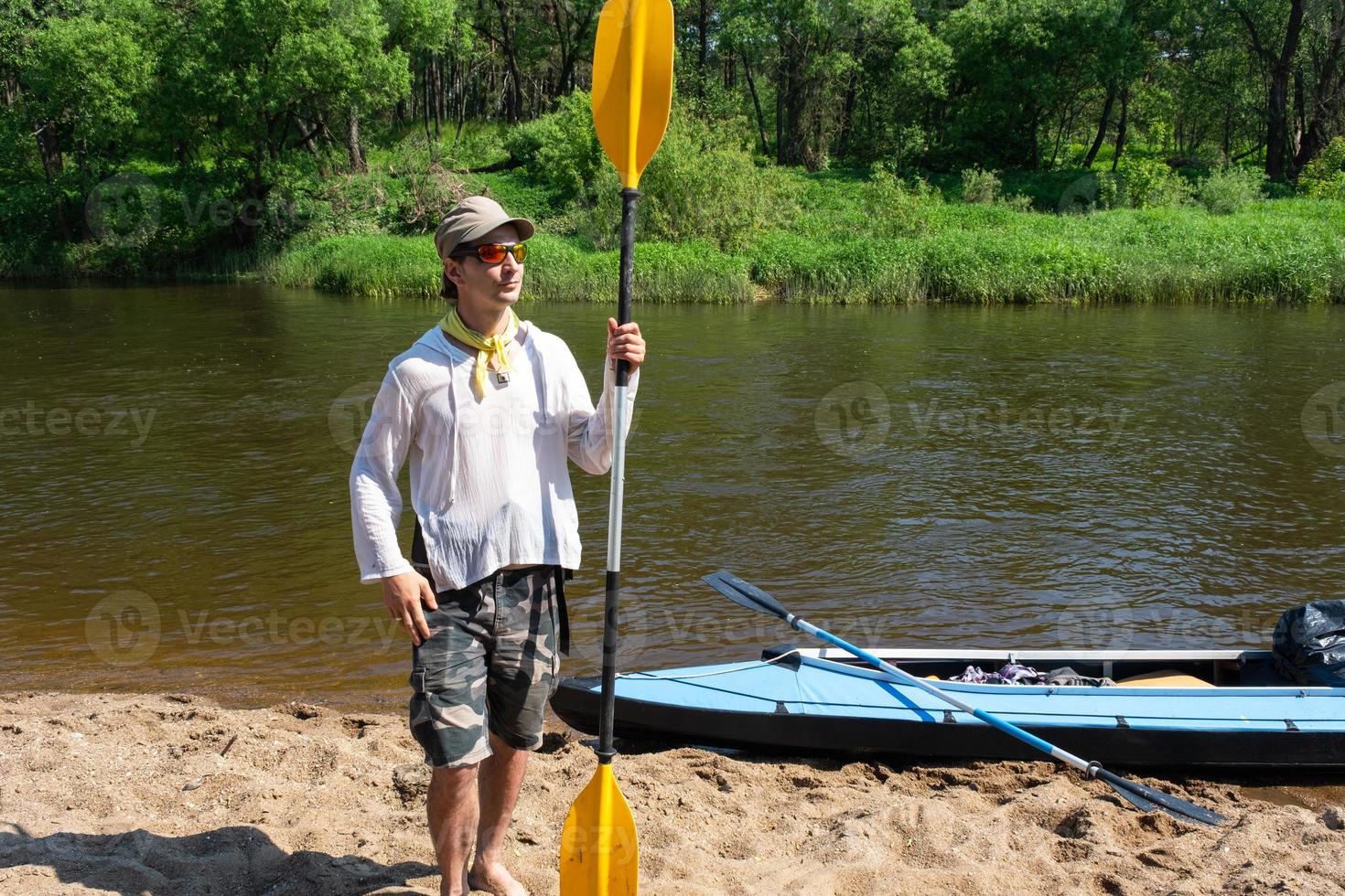 un hombre con un kayac paleta para canotaje soportes en el río banco. deporte agua caminata, un verano aventura. Respetuoso del medio ambiente y extremo turismo, activo y sano estilo de vida foto