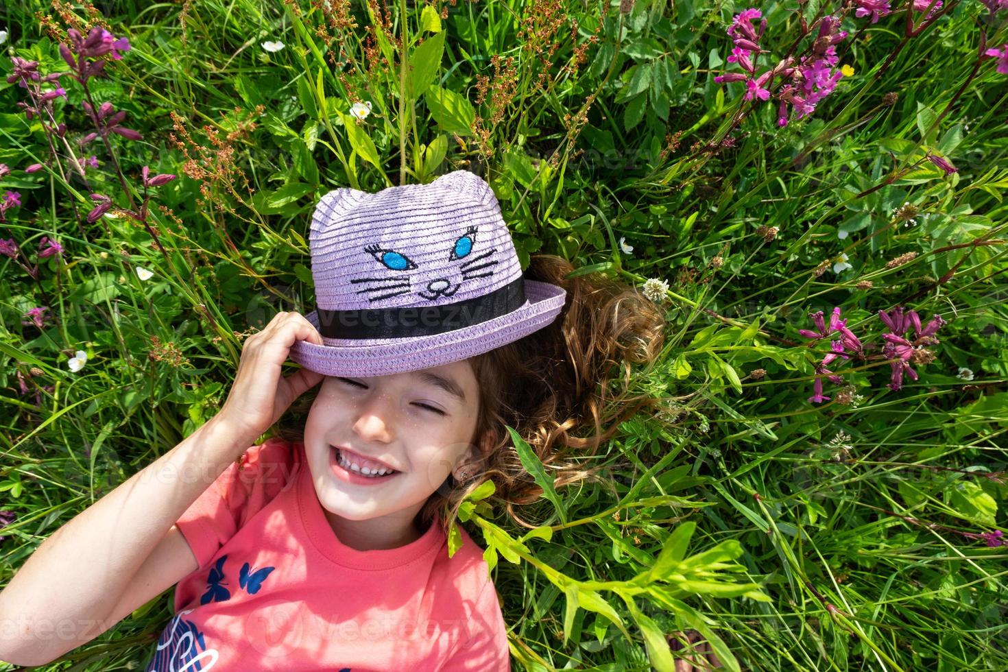 Portrait of a child in a hat with his face covered in summer lying in the grass and wildflowers. Hat with eyes and ears like a cat, summer time, freedom photo