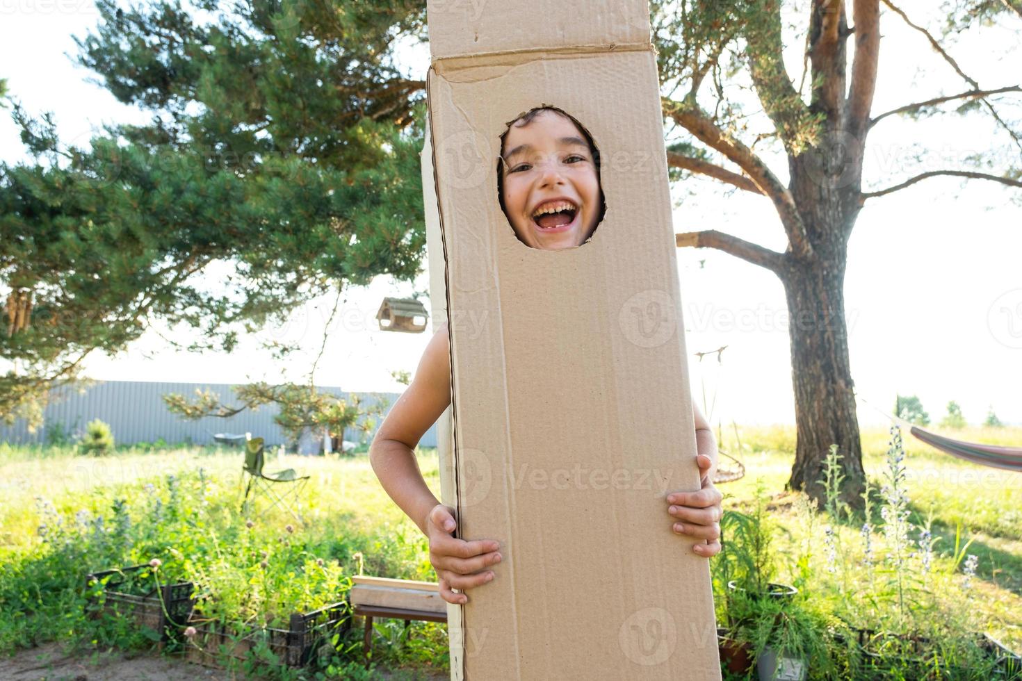 el niño es gracioso bailando y engañando alrededor en un caja disfraz - redondo separar para cara y manos. Moviente a un nuevo casa, para niños juegos desde improvisado medio, un traje con tu propio manos foto