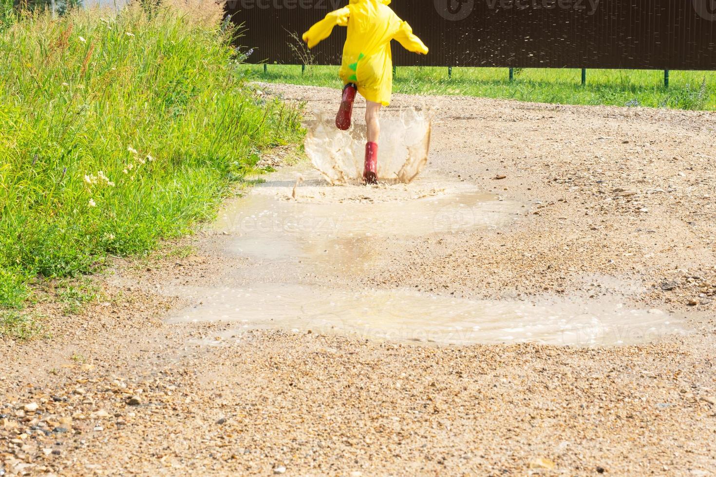 A girl in red rubber boots and a yellow raincoat runs through puddles after a rain in the village. Summer time, freedom, childhood photo