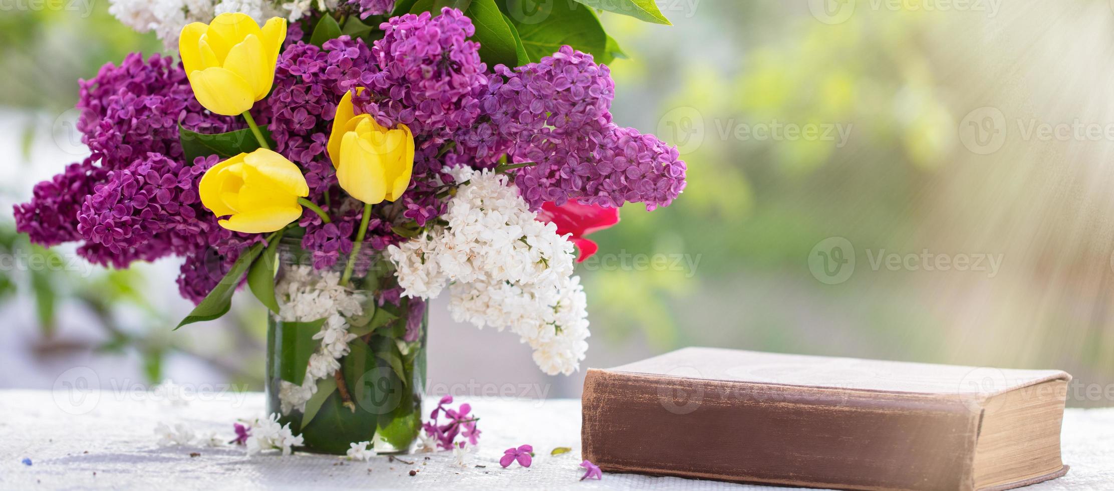 Spring background. Lilac and tulips in a glass jar on the table, also an old book. Beautiful flowers lit by the rays of the sun. photo