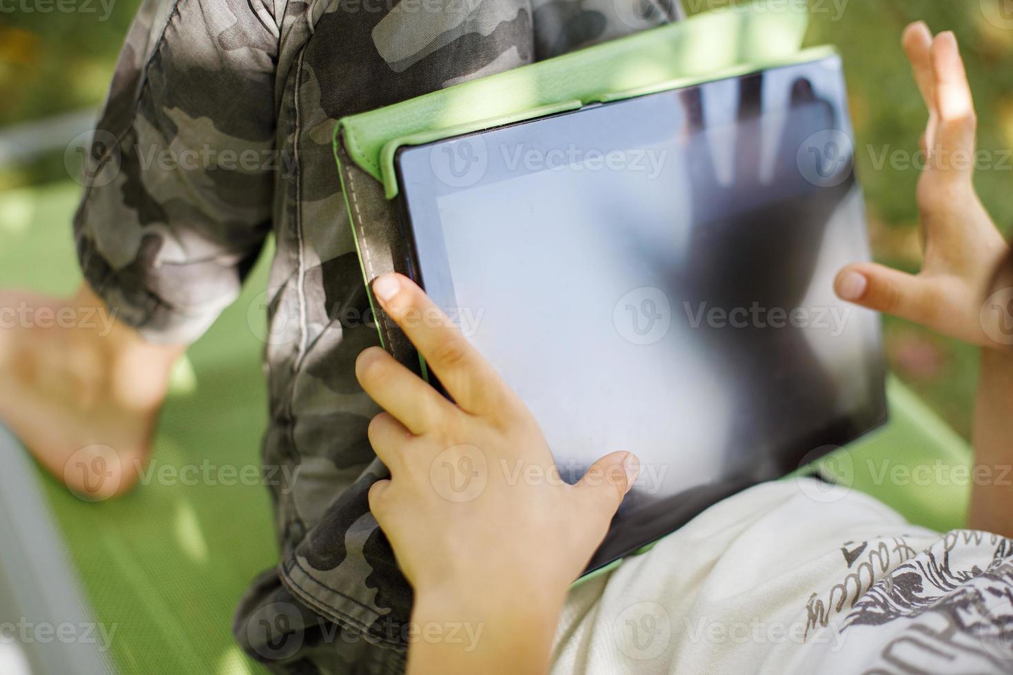 boy sitting with a tablet in the garden. the boy lies on a deck chair and plays on the tablet. child with a gadget photo