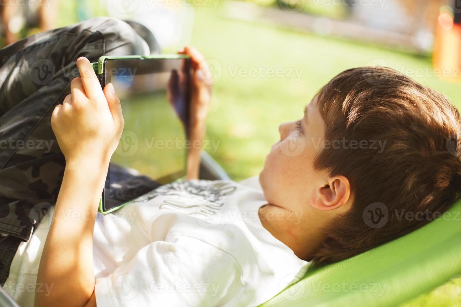 boy sitting with a tablet in the garden. the boy lies on a deck chair and plays on the tablet. child with a gadget photo