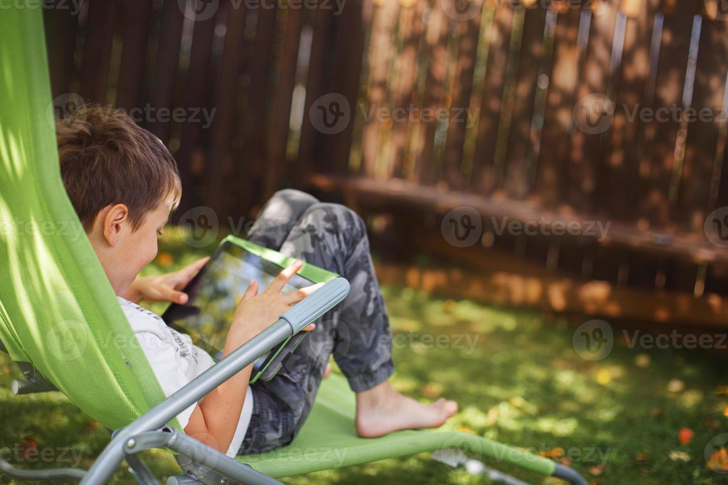 boy sitting with a tablet in the garden. the boy lies on a deck chair and plays on the tablet. child with a gadget photo