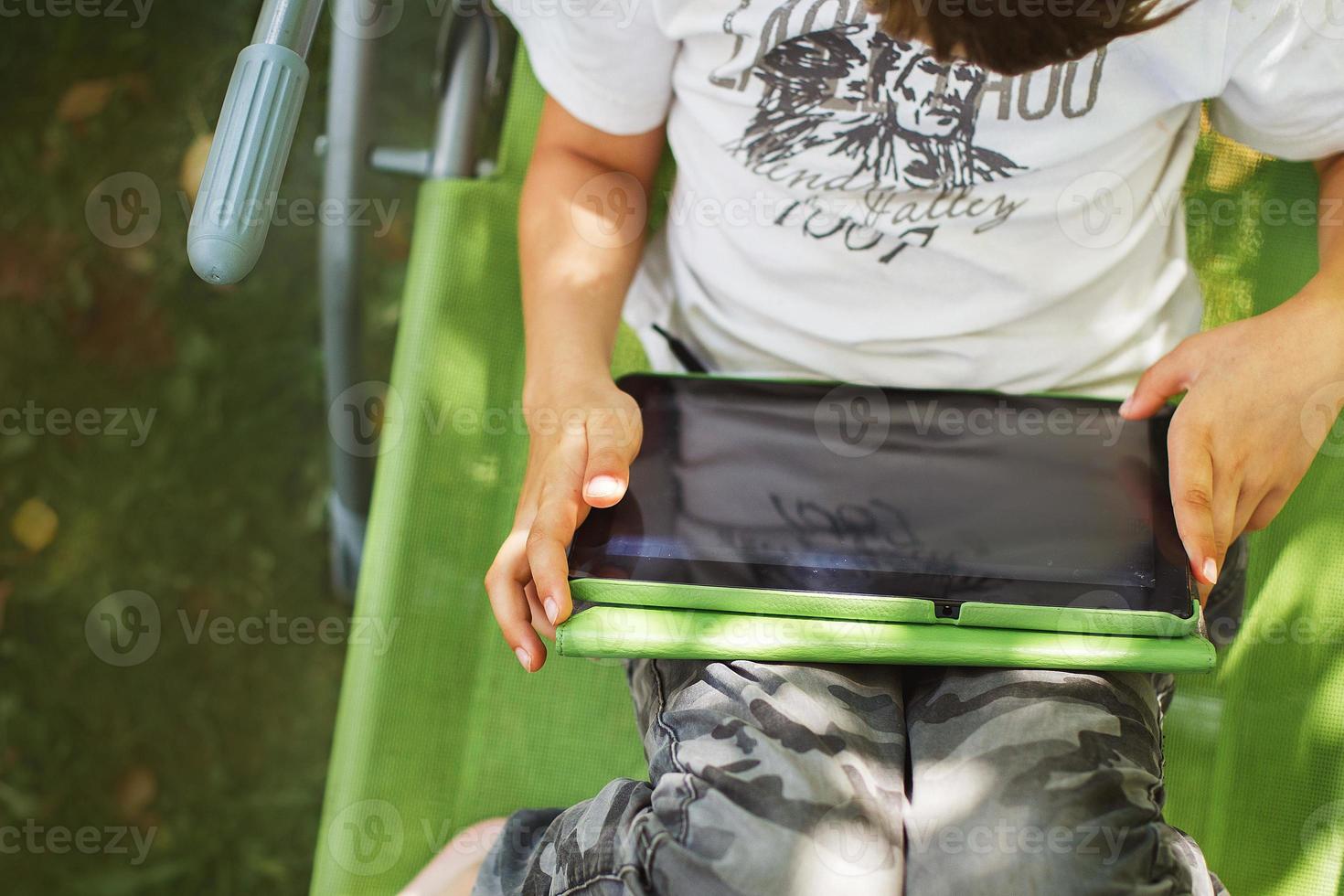 boy sitting with a tablet in the garden. the boy lies on a deck chair and plays on the tablet. child with a gadget photo