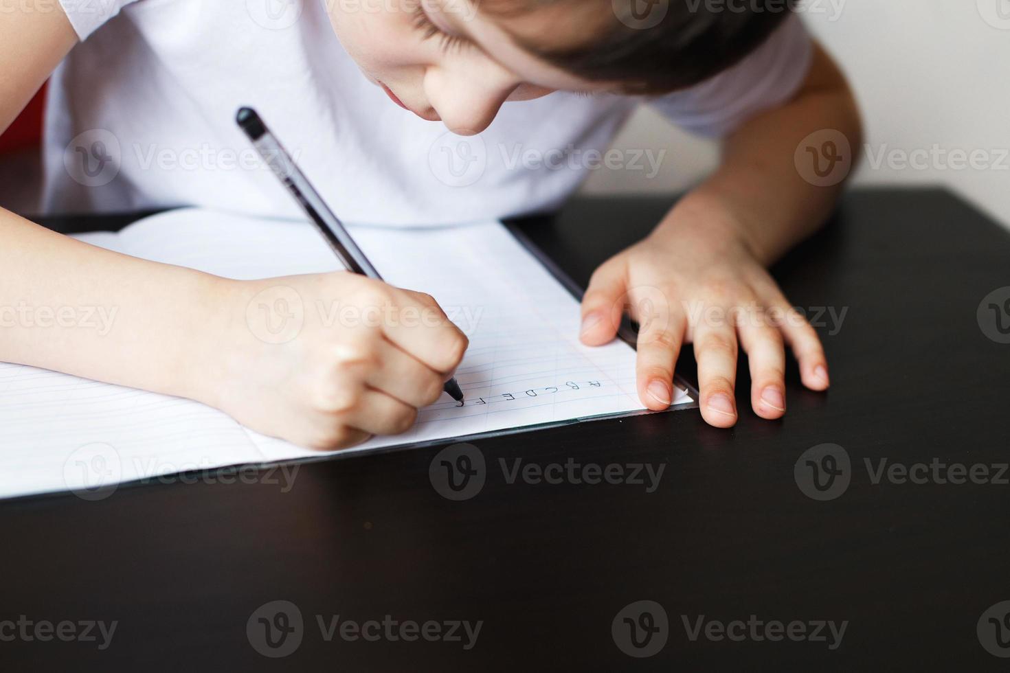 the boy sits at the table and writes in a notebook. child sits and does homework on a white background photo
