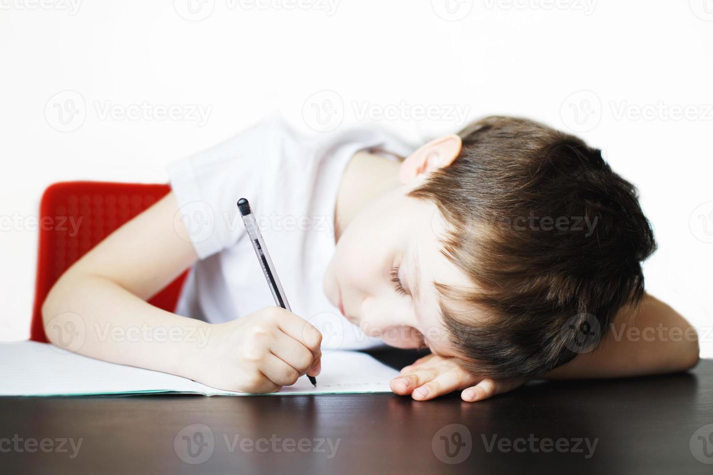 el chico se sienta a el mesa y escribe en un cuaderno. niño se sienta y lo hace deberes en un blanco antecedentes foto